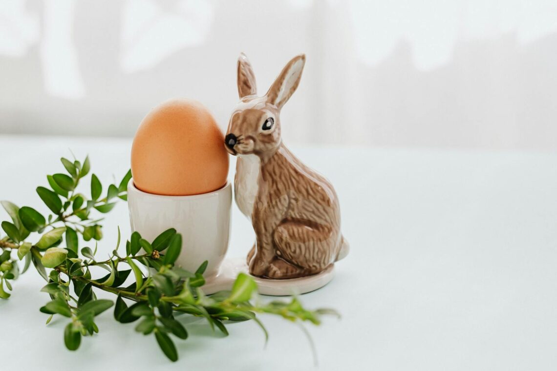 A festive Easter scene with a ceramic bunny, egg in holder, and greenery on a light background.
